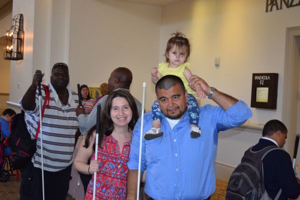 Blind parents pose at the NFB National Convention with their child sitting on the father's shoulders.