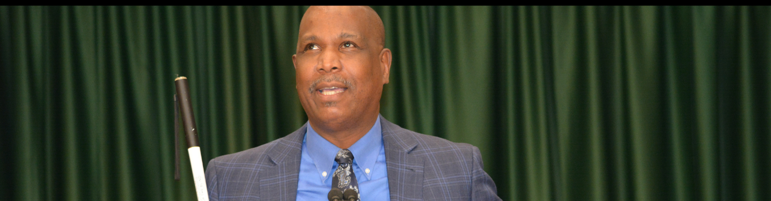 Anil Lewis speaks at podium, wearing a blue plaid suit and holding his white cane