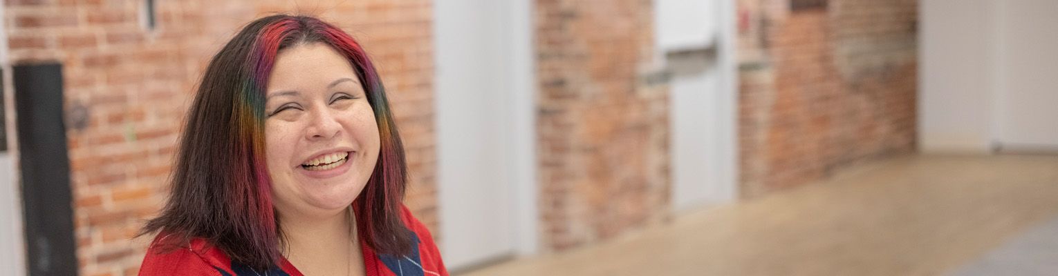 Headshot of Danielle McCann in front of brick wall. Ms. McCann has brown Shoulder length hair with multi-colored accents and is smiling.