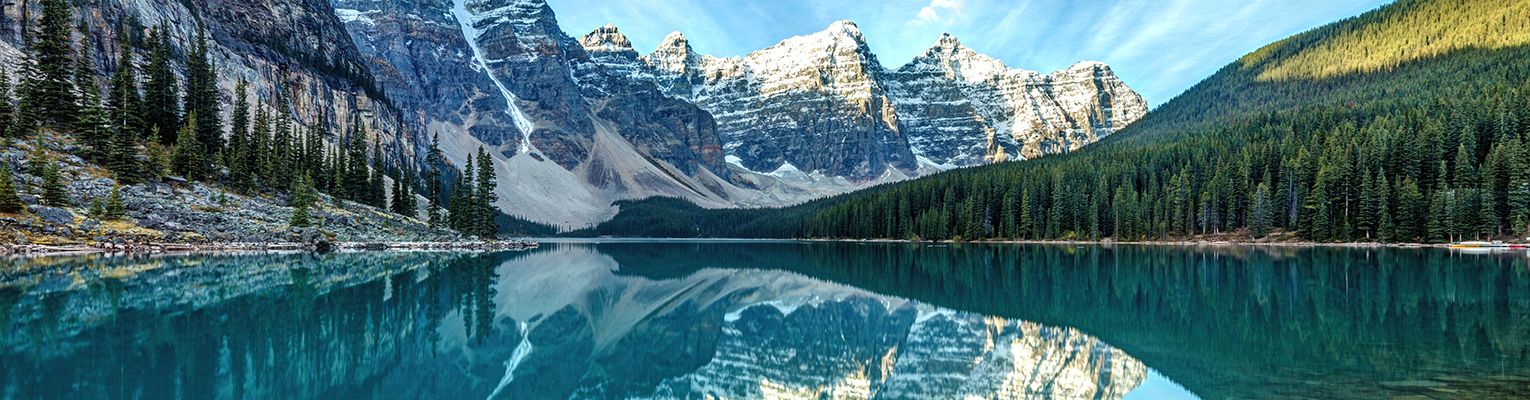 At the edge of a peaceful lake surrounded by snow-covered mountains on the left and dark green trees on the right.