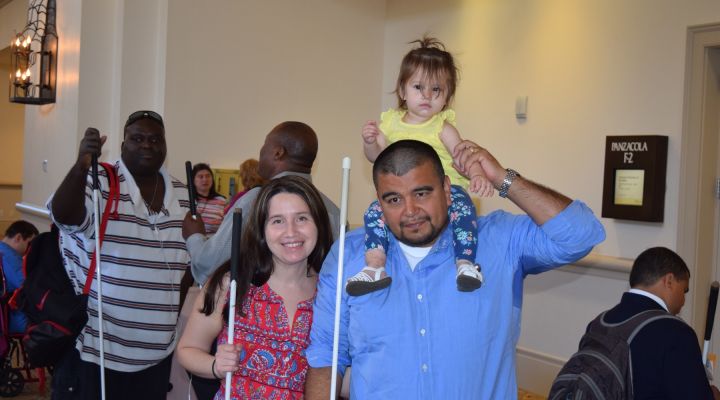 Blind parents pose at the NFB National Convention with their child sitting on the father's shoulders.